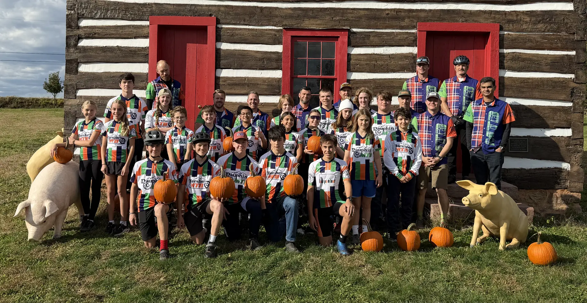 Hershey and Dauphin riders and coaches hold pumpkins in front of a log cabin at Punktoberfest 2025.